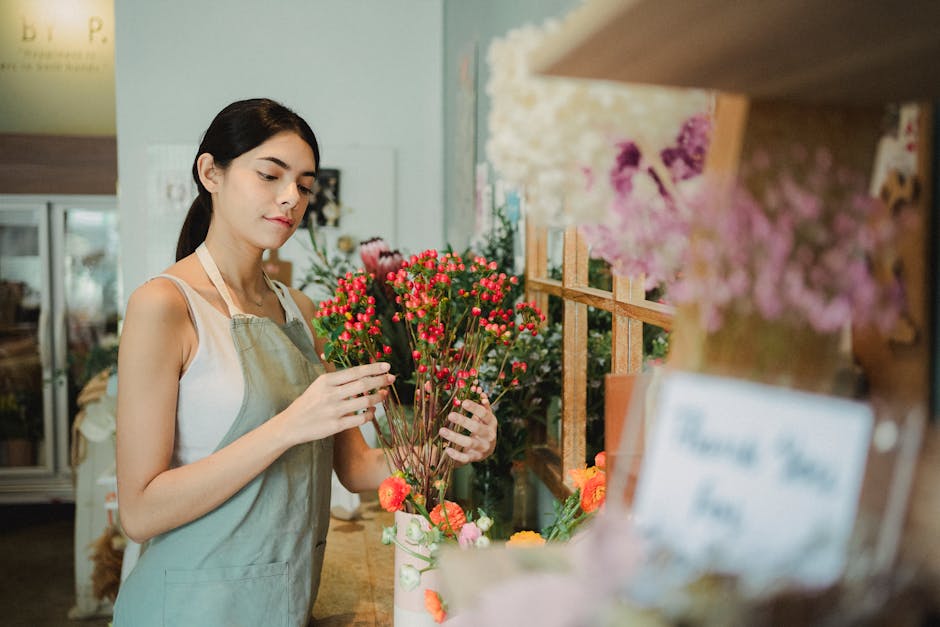 A young female florist with dark hair pulled back, wearing a white sleeveless top and a light-colored apron, arranging a bouquet of small, vibrant pink flowers with green stems and buds. She is standi