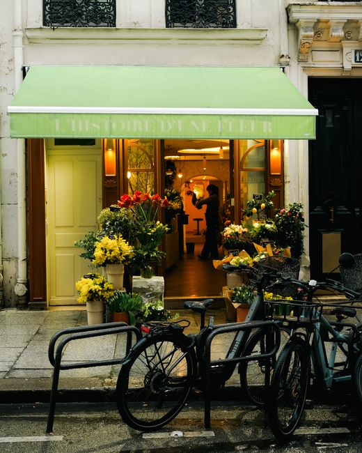 The entrance of a florist shop with a green awning inscribed 'HISTOIRE DU FLEUR' in white, displaying a variety of fresh flower arrangements in buckets and vases, including bright yellow chrysanthemum