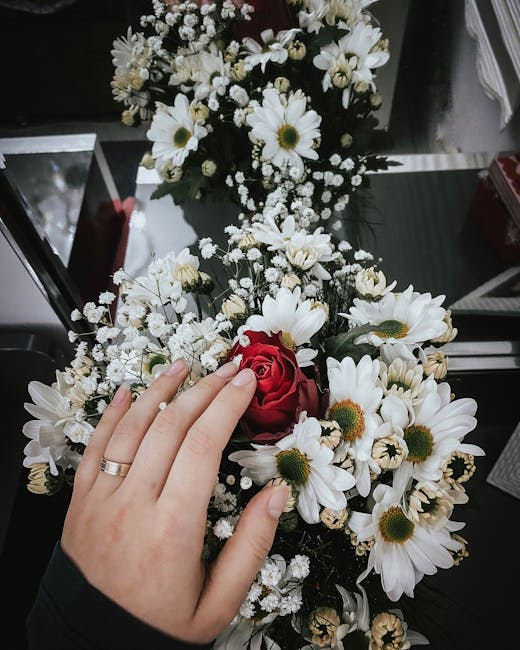 A close-up of a floral arrangement featuring a mix of white daisy-like flowers with yellow centers, small white baby's breath, and a single deep red rose, held by a person with a light skin tone and a