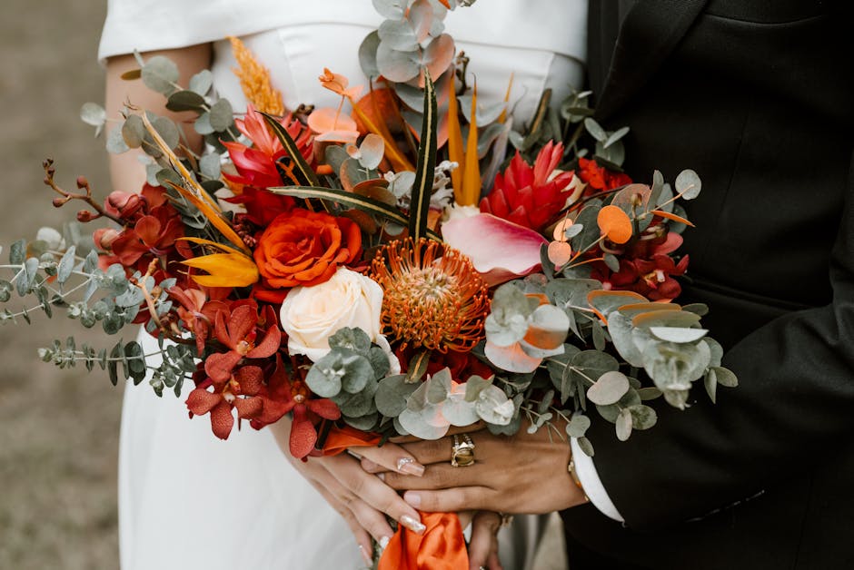 A wedding bouquet held by a bride and groom, featuring a vibrant arrangement of flowers including orange roses, white calla lilies, red and pink proteas, and lush greenery like eucalyptus with rounded