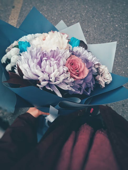 A hand holding a bouquet of fresh flowers wrapped in blue and white paper, featuring a pink rose, large purple and white chrysanthemums, and bright blue flowers. The arrangement is neatly assembled wi