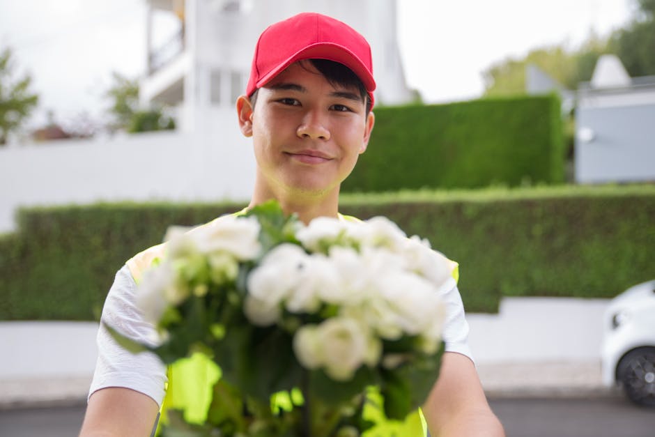 A delivery person wearing a red cap and a reflective vest is standing outdoors, holding a bouquet of fresh white flowers with green foliage. The flowers appear to be in good condition with soft petals