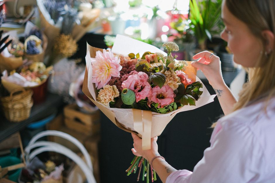 A woman holding a vibrant bouquet of fresh flowers, including large pink dahlias, pale pink and cream roses, dark green foliage, and small clusters of white baby's breath, wrapped in light brown paper